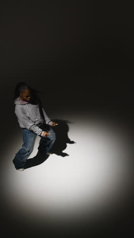 Vertical-Video-Full-Length-Studio-Portrait-Shot-Of-Young-Woman-Wearing-Hoodie-Dancing-In-Spotlight-With-Shadow-1